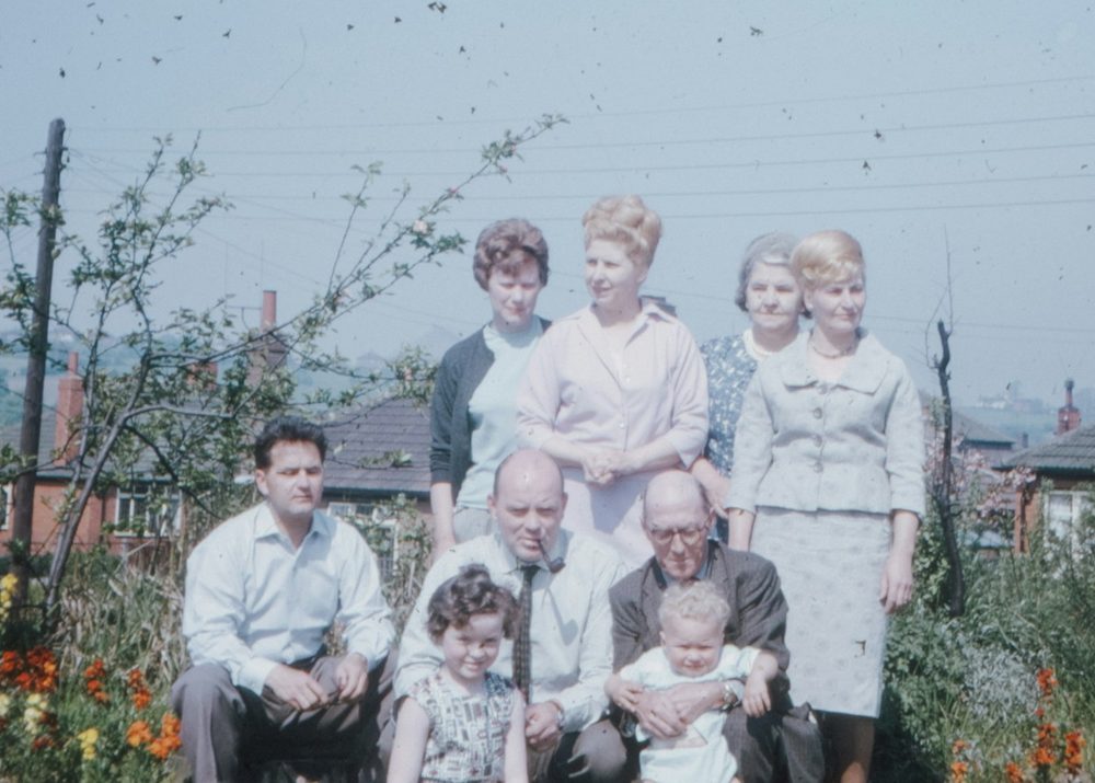 A faded 1960s colour photograph of a family in a garden, with people nobody can yet name